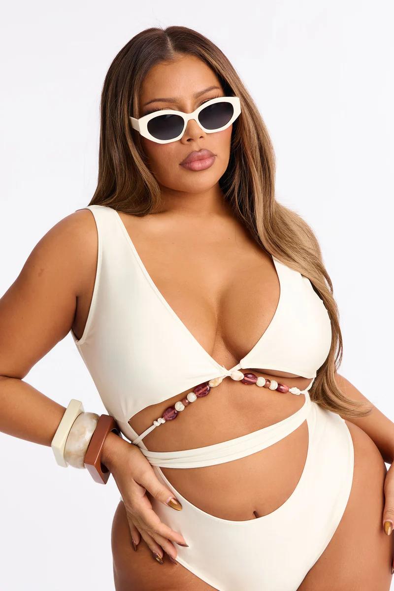 Plus-size woman in cream one-piece swimsuit with cutouts, purple/cream beaded detail, white sunglasses, and bangles. Discover new beachwear.