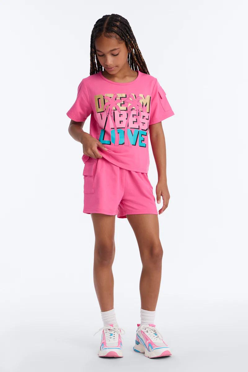 Girl in a pink 'DREAM VIBES LIVE' tee with cargo shorts and colorful sneakers. Ideal girls' summer collection item.
