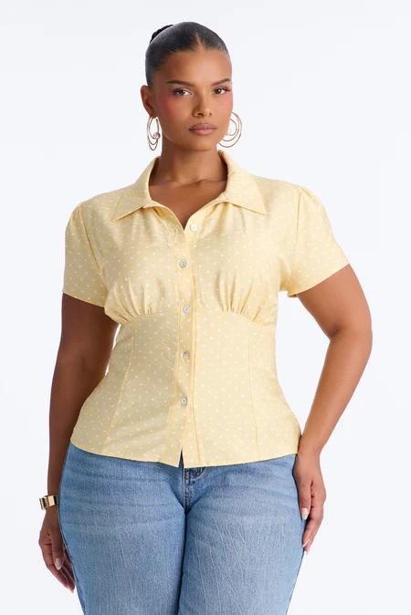 Woman in a collared, short-sleeved yellow shirt with white polka dots, fitted waist, and blue jeans. She wears gold hoop earrings.