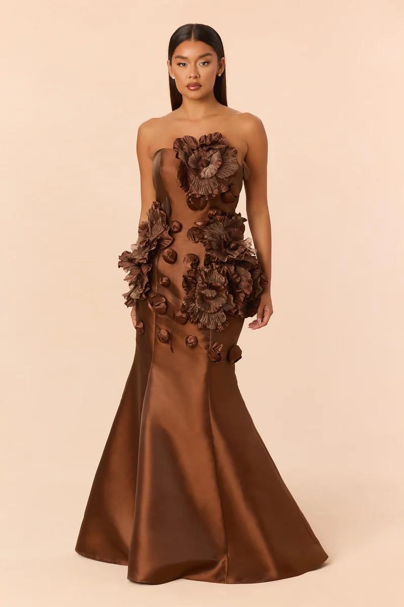 Model in an elegant brown strapless mermaid gown with large 3D floral appliques, promoting a luxury formal evening wear collection.