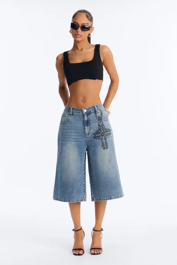 Full-body shot of a woman in a black square-neck crop top, light blue denim culottes with a dark gray embellished cross, and black strappy heels, wearing sunglasses.