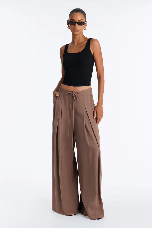 Woman posing in a sleek black crop tank top and brown pleated wide-leg linen pants, showcasing a trendy summer fashion collection.