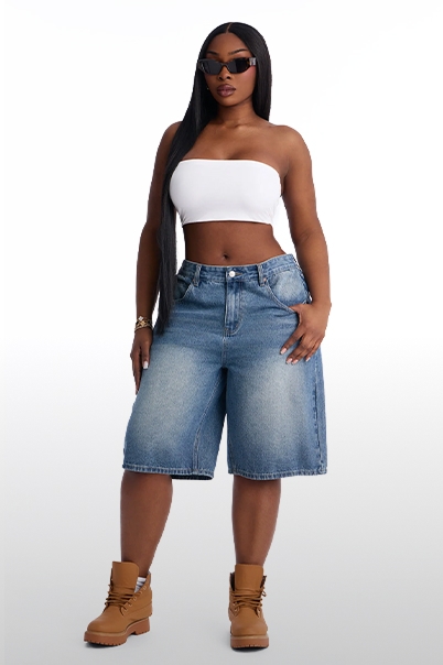 Mujer con top blanco sin tirantes, bermudas de mezclilla azul desteñida, botas marrones y gafas, promocionando moda casual chic femenina.