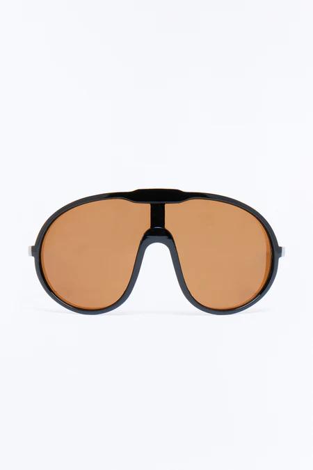 Gafas de sol negras tipo escudo, de gran tamaño, con lentes marrones cálidas y puente central, centradas precisamente sobre fondo blanco.