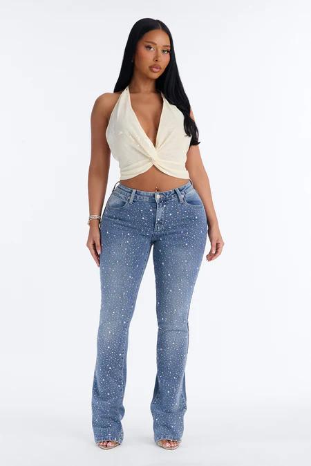 Woman in pale yellow twisted halter crop top and blue flared jeans with sparkling white rhinestones, standing against a white background.