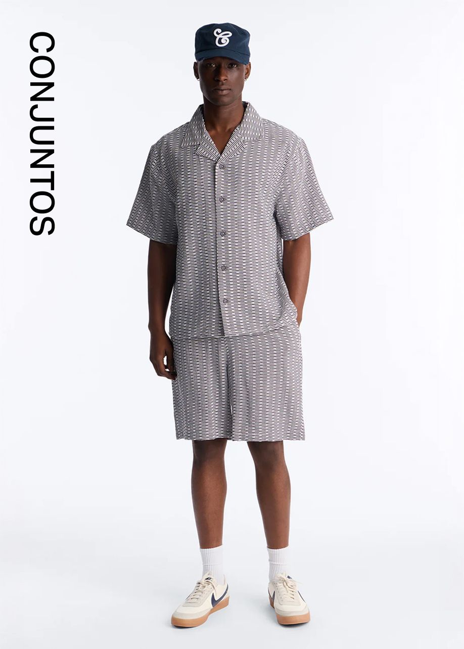 Hombre con conjunto de camisa y shorts estampados gris-blanco y gorra azul marino. Descubre conjuntos a juego de moda masculina de verano.