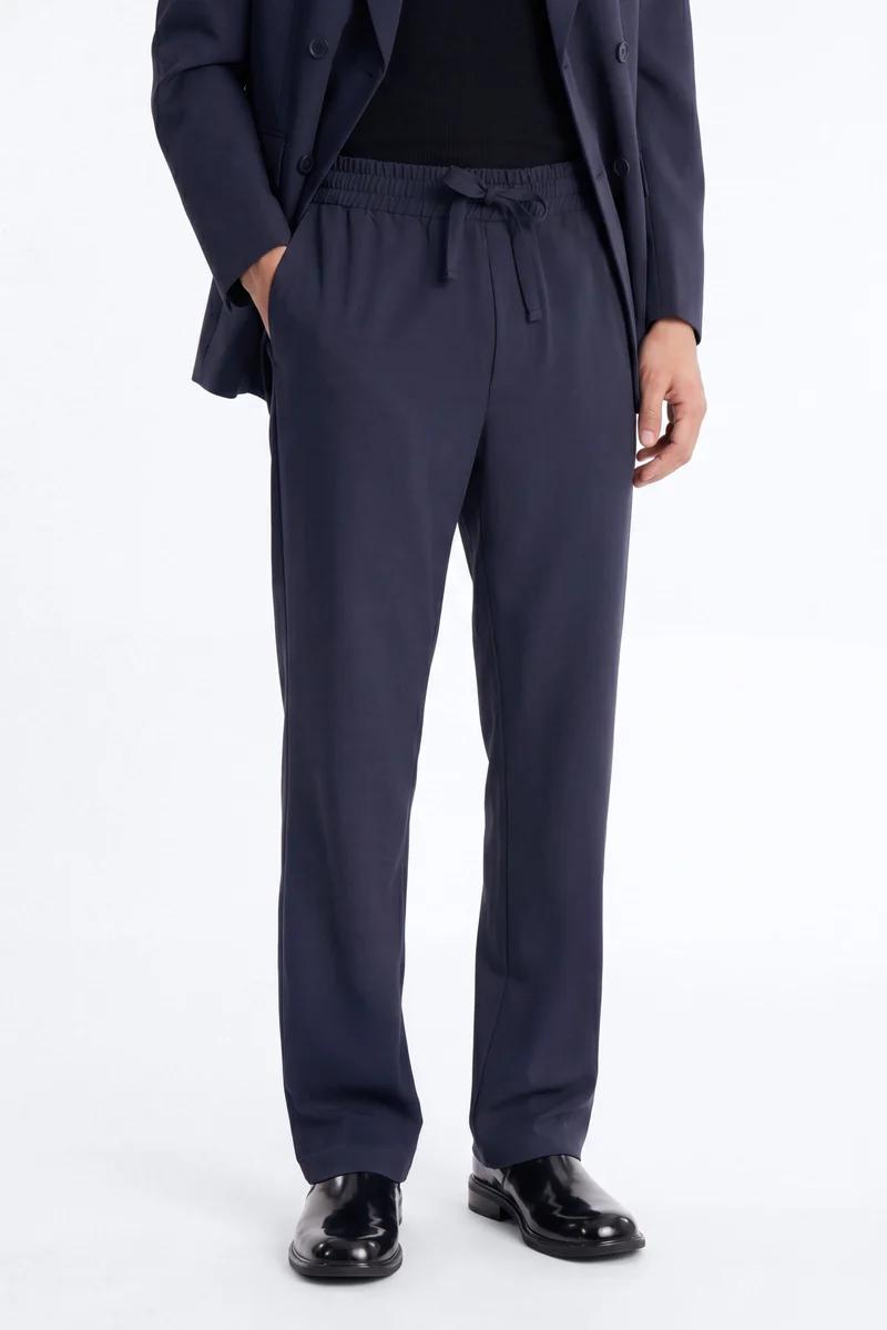 Hombre con elegantes pantalones azul marino con cordón, blazer oscuro y zapatos negros, destacando moda masculina cómoda e inteligente.