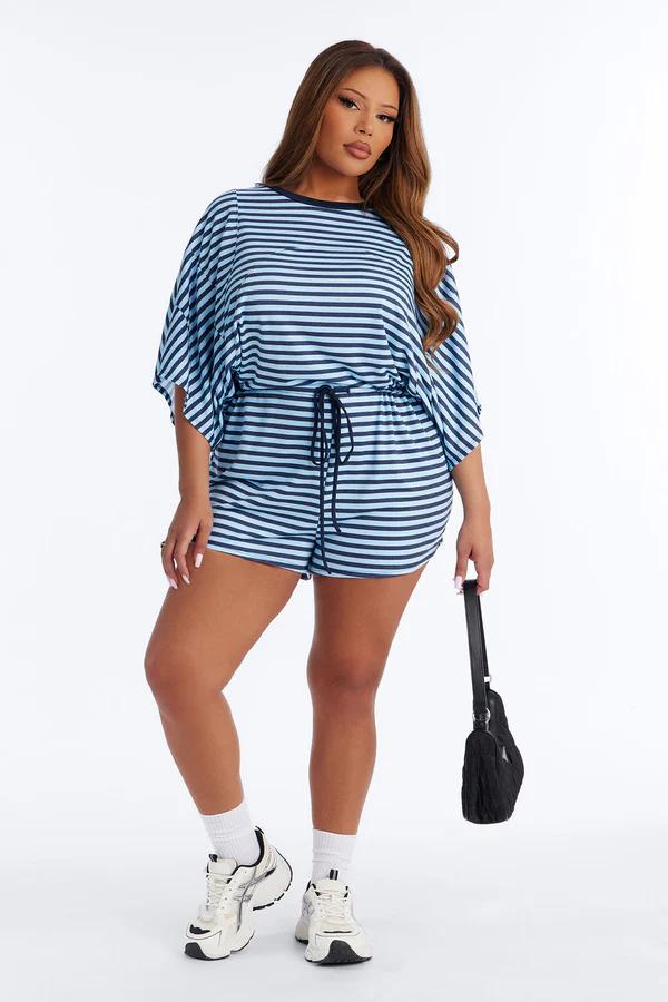 Mujer modela mono de rayas azul y blanco de tallas grandes con cintura fruncida, zapatillas blancas y bolso negro.