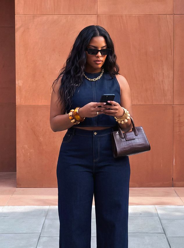 Stylish woman in dark denim crop top and wide-leg jeans, gold accessories, brown bag, scrolling phone against a terracotta wall. Shop the look!