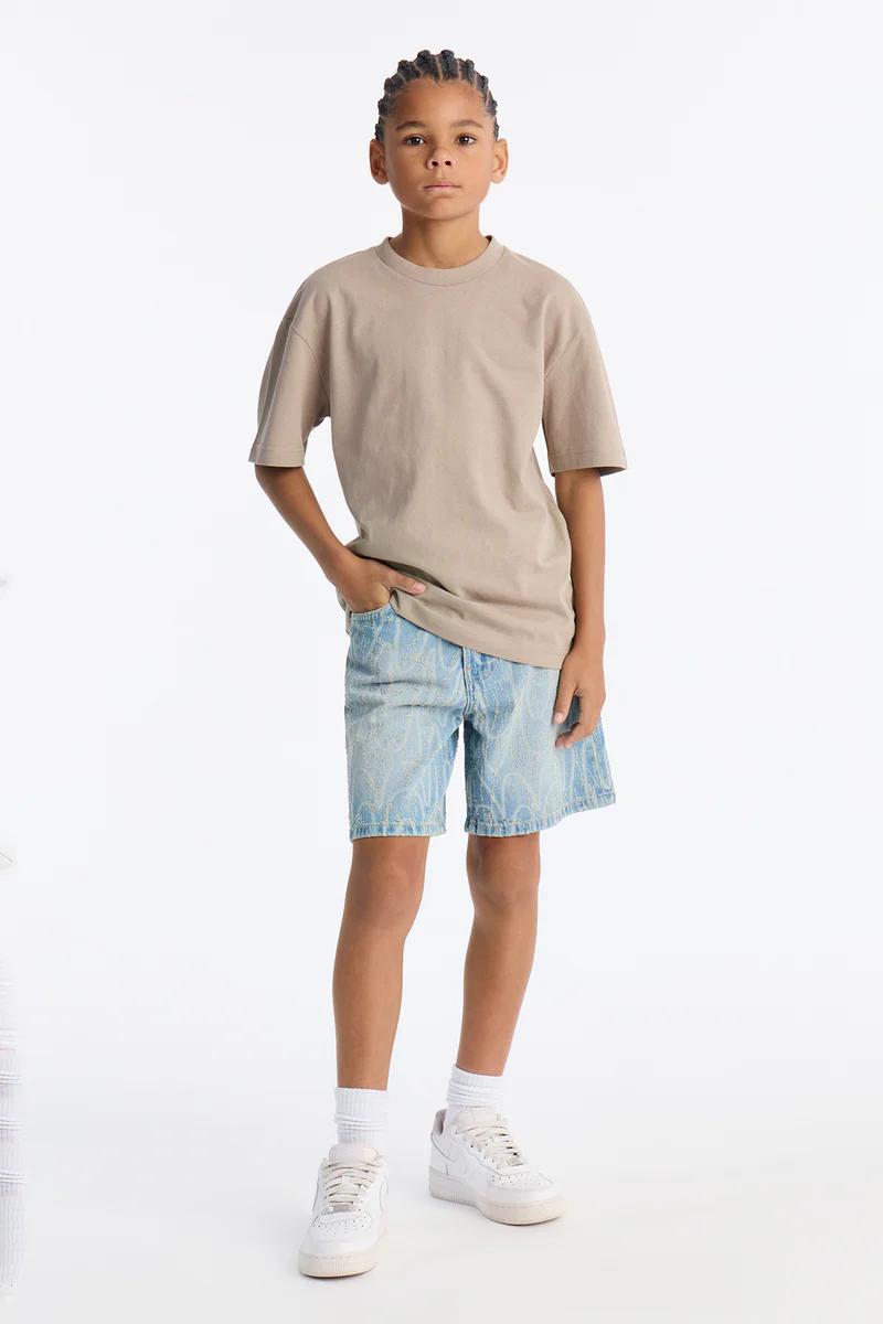 Niño joven con camiseta oversize beige, shorts vaqueros con patrón azul claro y zapatillas blancas. Mostrando ropa casual de verano para niños.