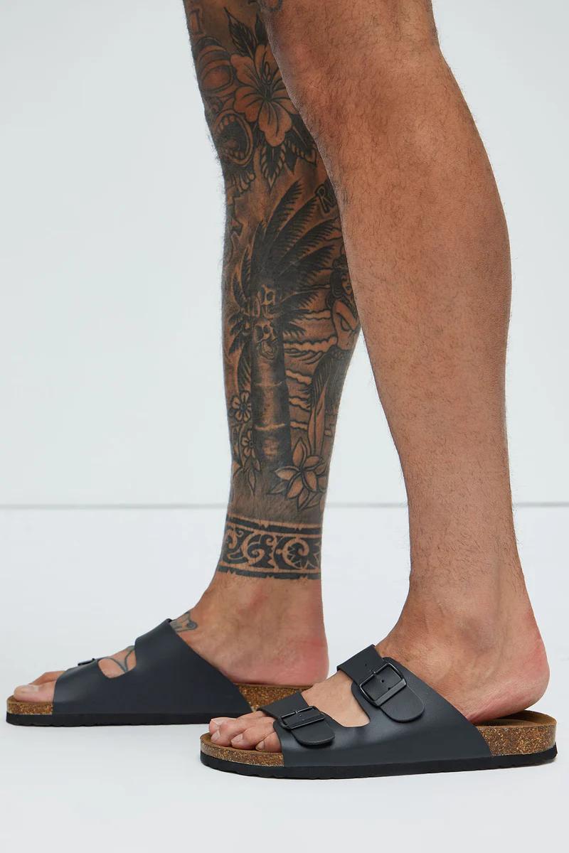 Man's legs, one with a tropical tattoo, display black adjustable buckle sandals with cork footbeds for comfortable summer style.