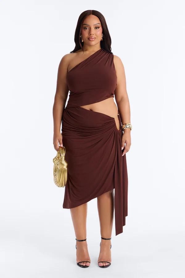 Mujer con vestido midi marrón de un hombro fruncido con aberturas, sosteniendo un bolso dorado. Elegante vestido de fiesta talla grande para un look chic.