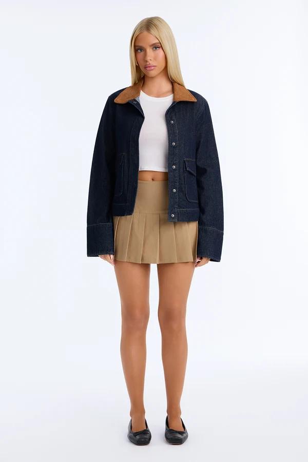 Blonde woman wearing a dark denim jacket with brown collar, white crop top, and tan pleated mini skirt for a trendy casual fall fashion style.
