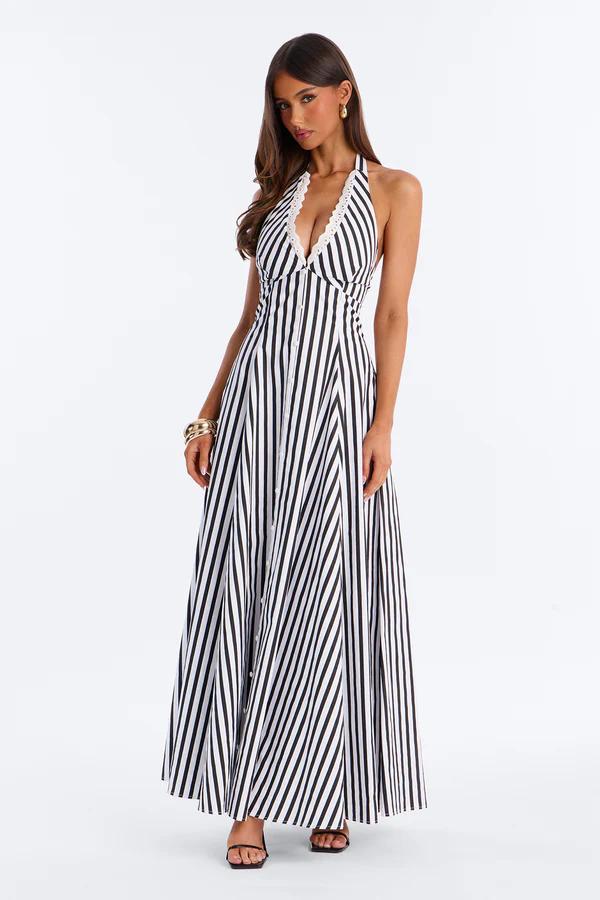 Woman in a square-neck, white maxi dress with blue floral print, bow straps, and a lace hem, holding a straw clutch.
