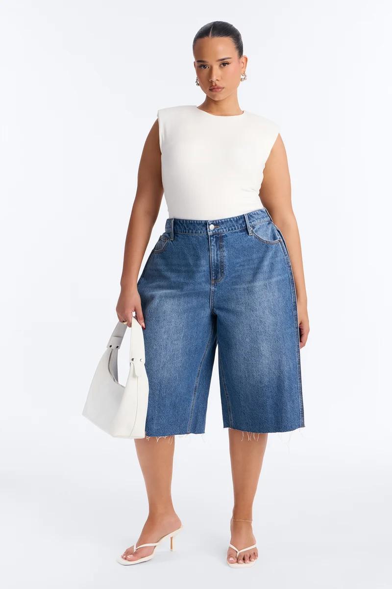 Plus size woman in a white sleeveless top, high-waisted blue denim bermuda shorts, white heels, and holding a white handbag.