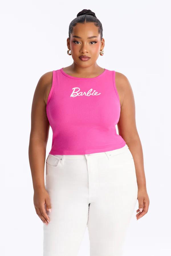 Woman in a pink ribbed Barbie tank top and white jeans, standing confidently against a white background. Dark hair is in a bun.