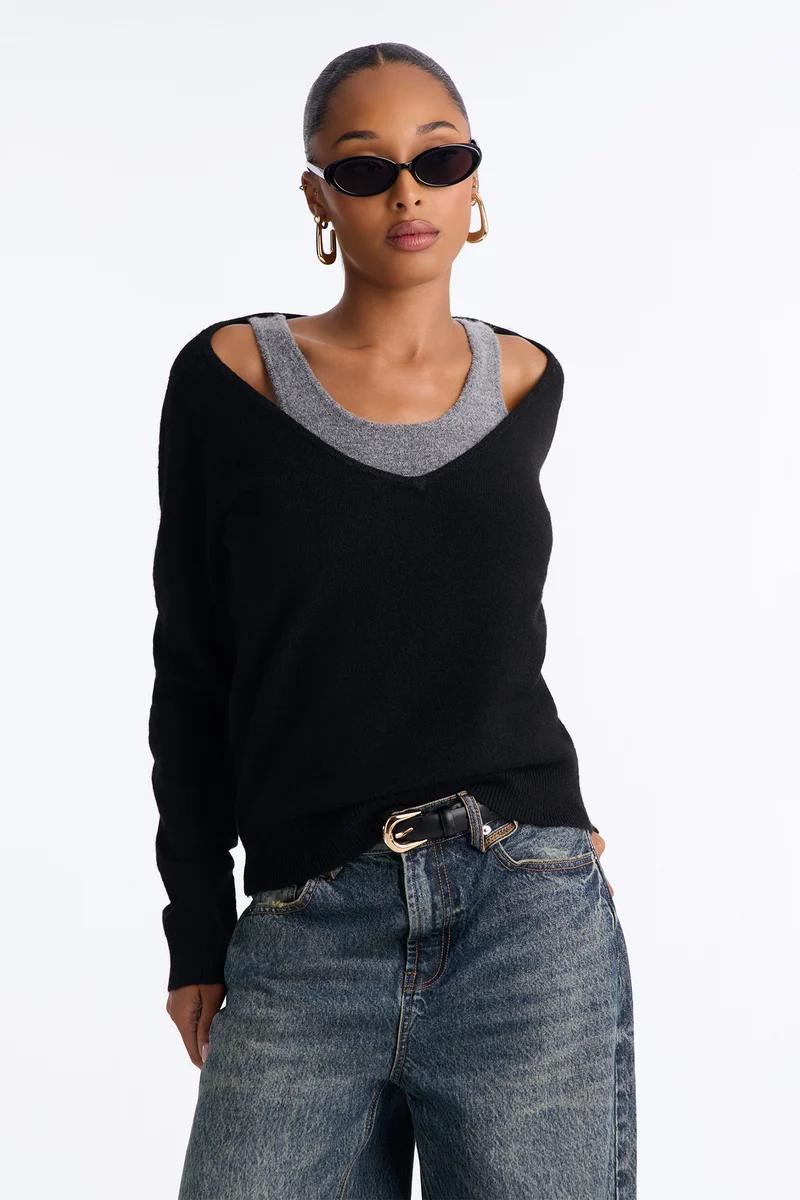 Mujer con suéter negro de hombros caídos, camiseta de tirantes gris y vaqueros azules.