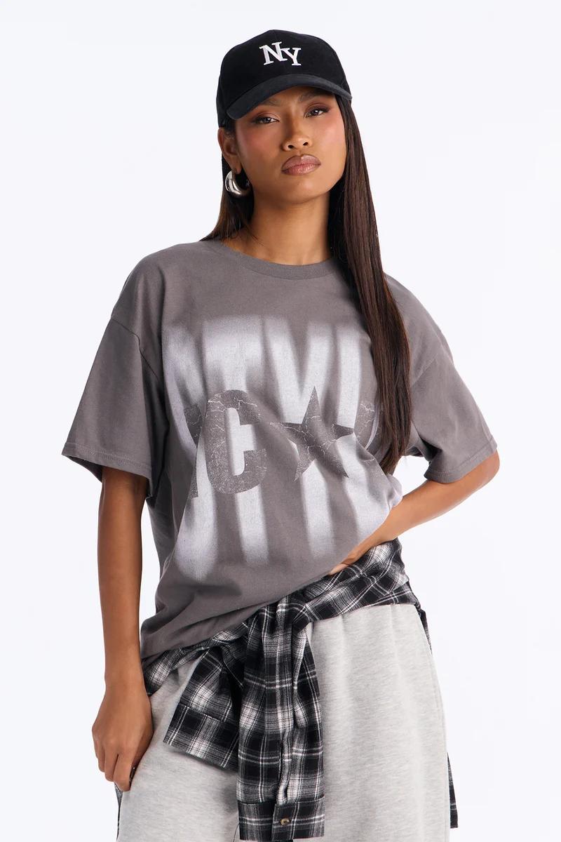 Modelo con camiseta gráfica gris, gorra negra de NY y camisa de cuadros en la cintura.