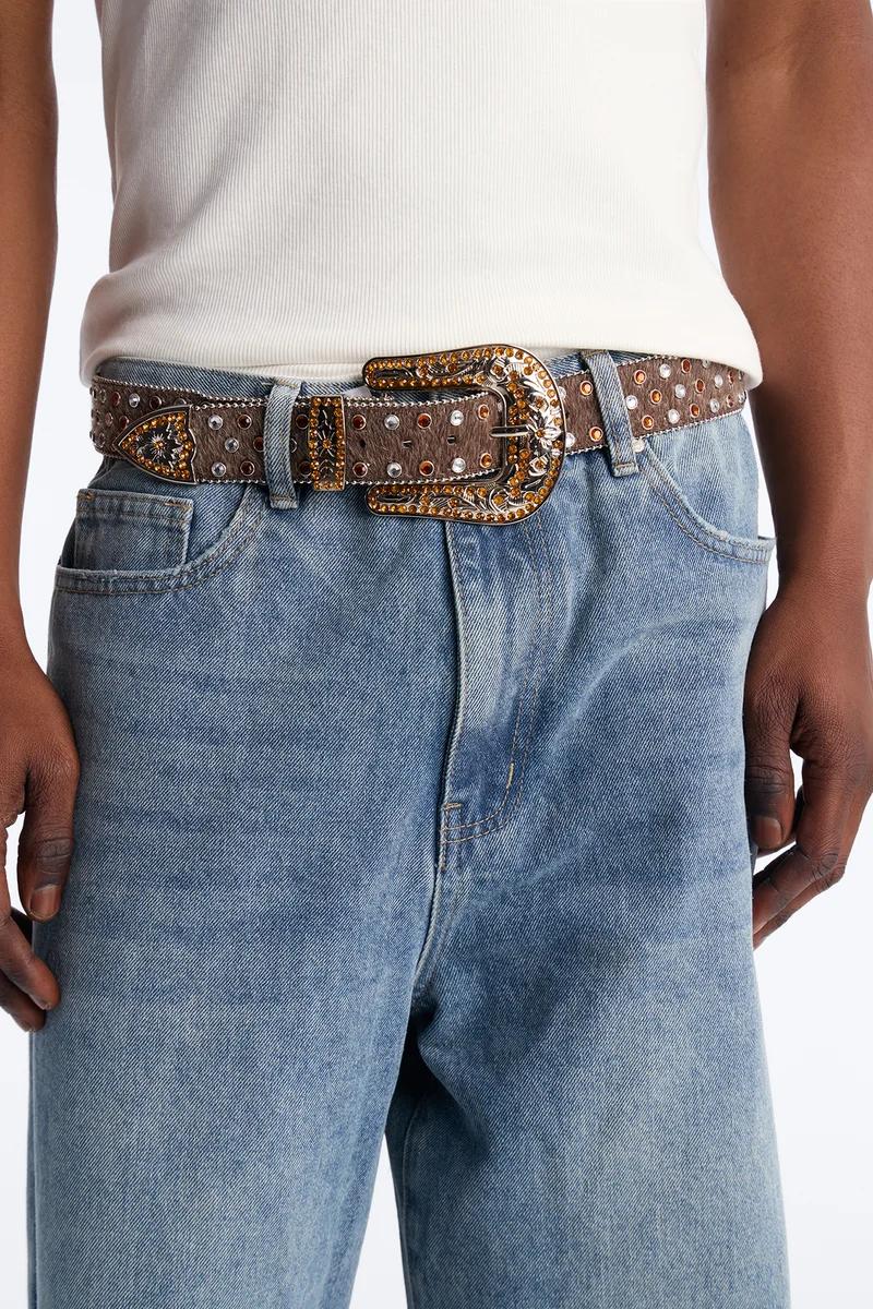 Brown textured western belt, studded with clear and amber rhinestones, showcasing an ornate silver and gold statement buckle on blue jeans.