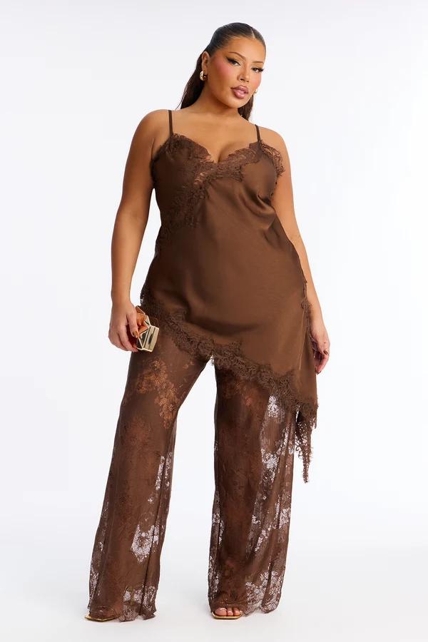 Plus size model in a chic brown satin asymmetric camisole and sheer lace pants set, showcasing a new fashion collection against a white backdrop.