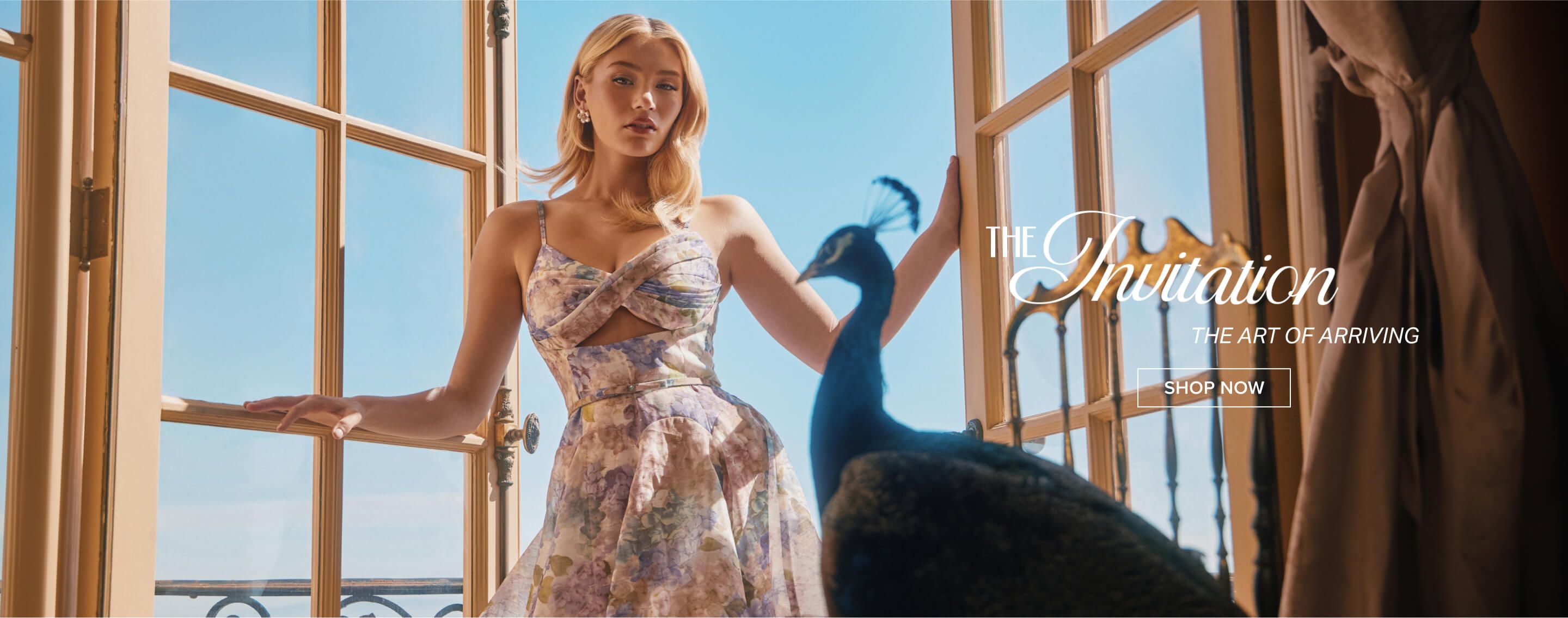 Blonde model in a pastel floral mini dress stands in a doorway with a peacock, promoting the new spring collection. Shop women's fashion now.
