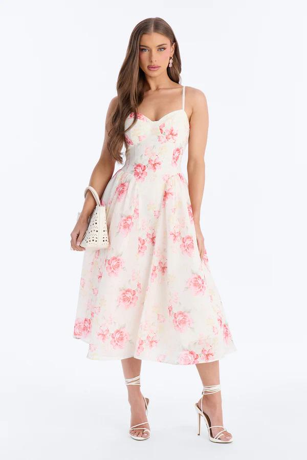 Woman in a square-neck, white maxi dress with blue floral print, bow straps, and a lace hem, holding a straw clutch.