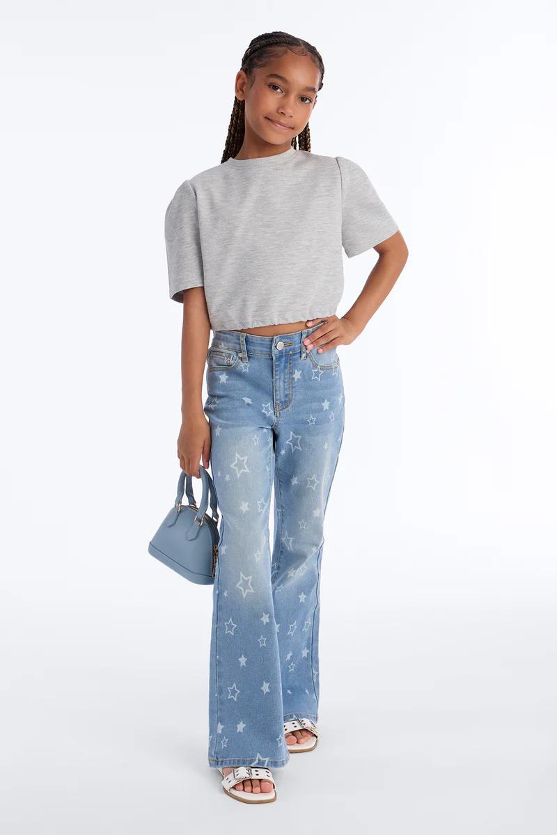 Girl in grey puff-sleeve crop top, star print flared jeans, blue bag, and white sandals. Trendy kids' fashion.