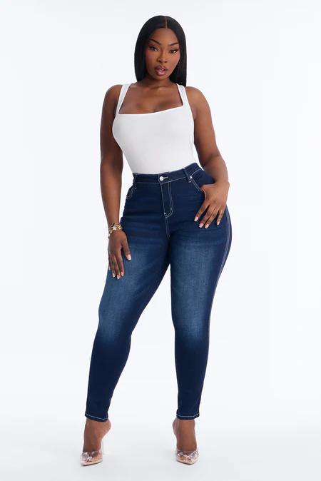 Mujer con top blanco de cuello cuadrado y jeans ajustados oscuros de talle alto con costuras claras, de pie sobre fondo blanco.