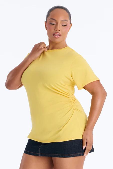 Mujer con top amarillo caléndula fruncido de manga corta y minifalda de mezclilla oscura, mano en el cuello, mirando hacia abajo.