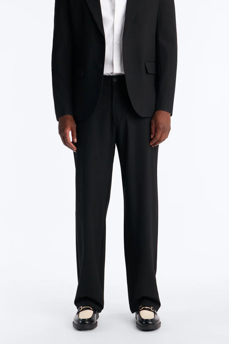 Man models a sleek black suit, white shirt, and black tailored trousers with classic gold-bit loafers. Ideal men's formal fashion.