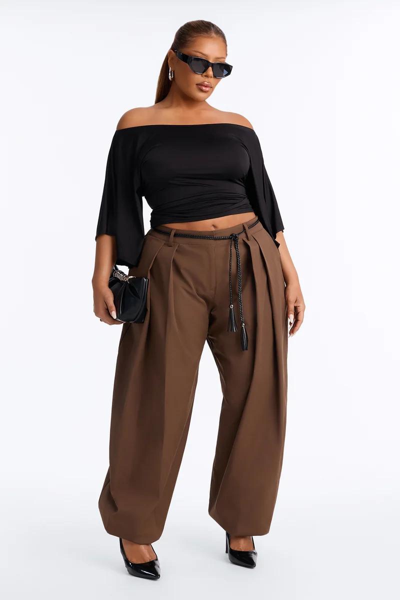 Modelo con top negro de hombros descubiertos y pantalones marrones de pinzas anchos, promocionando una colección de moda de tallas grandes.