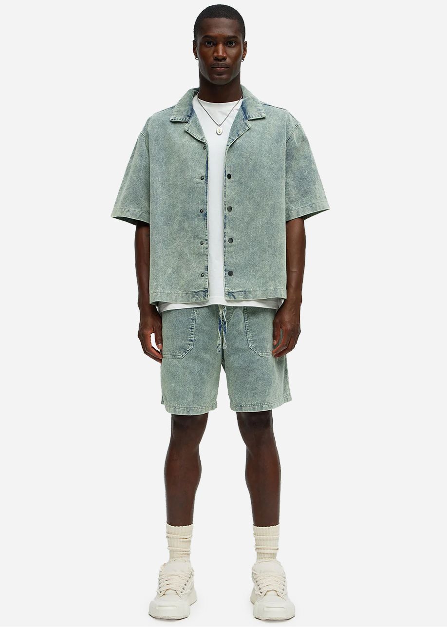 Hombre modela conjunto denim acid wash celeste: camisa manga corta, camiseta blanca, shorts con cordón. Descubre moda casual de verano.