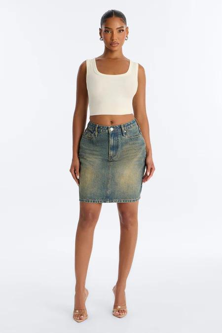 Mujer con top corto crema de cuello cuadrado, minifalda vaquera azul desgastada y mules de tacón transparentes, posando sobre fondo blanco.