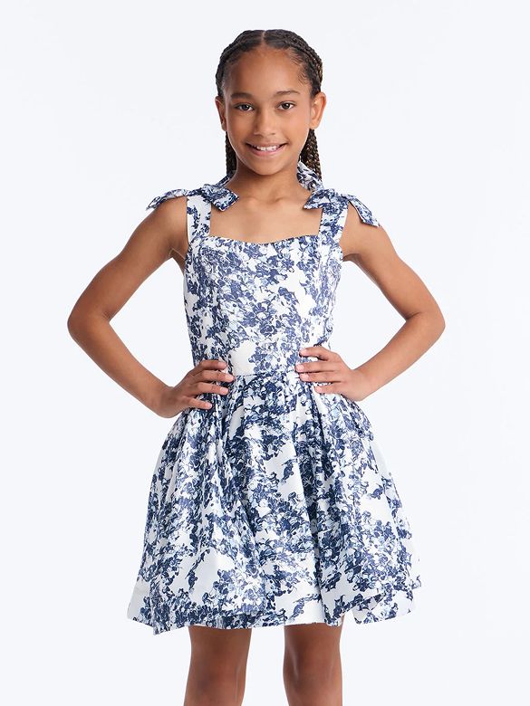 Smiling girl in a white and blue floral print dress with shoulder ties, hands on hips. A charming and stylish girls' party dress.