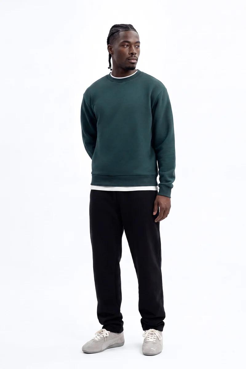 Man wears dark green crewneck sweatshirt, black joggers, and grey sneakers, showcasing stylish, comfortable men's casual wear.