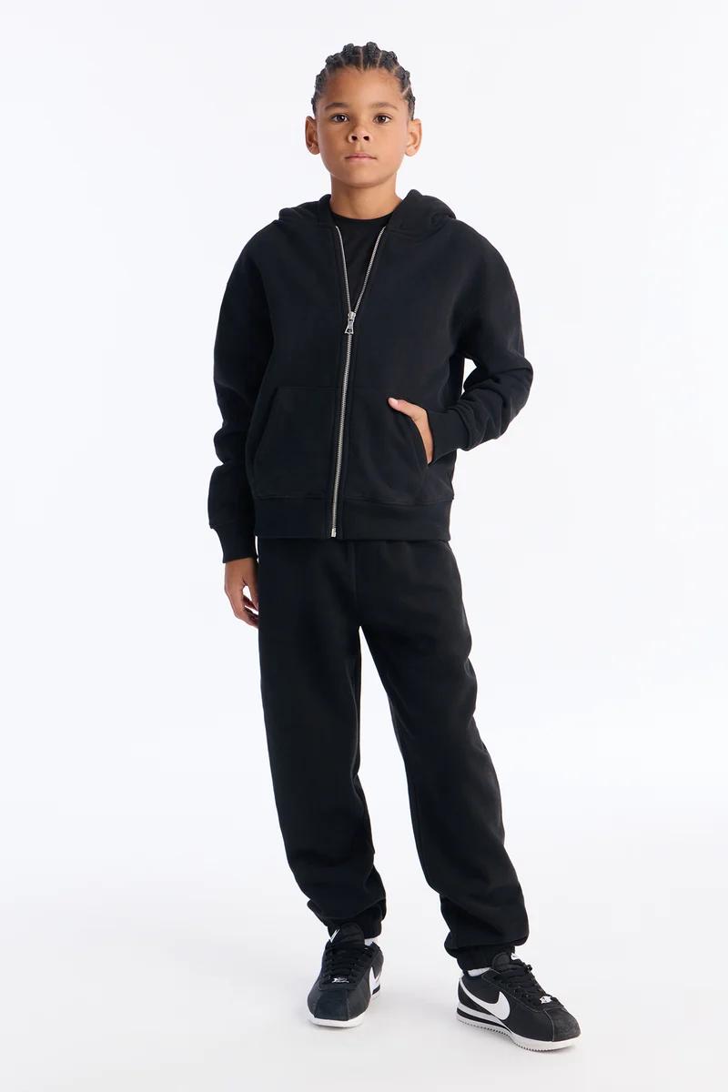 Young boy wearing a black zip-up hoodie, matching sweatpants, and black sneakers.