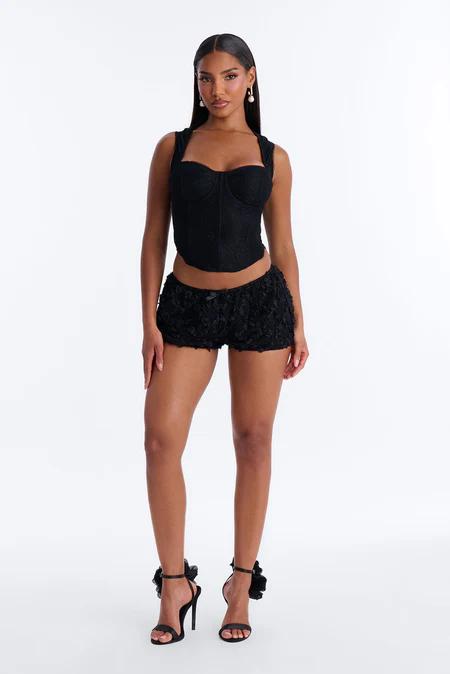 Woman in black lace corset top and textured shorts, standing on a white background. She wears black heels with ruffled ankle straps.