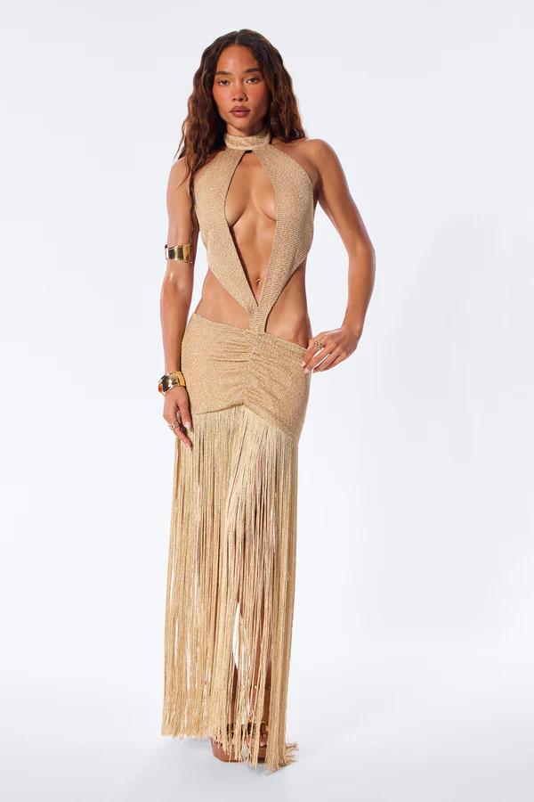 Woman posing in a two-piece brown and cream striped knit outfit with tiered fringe, a waist cutout, gold arm cuff, and braided clutch bag.