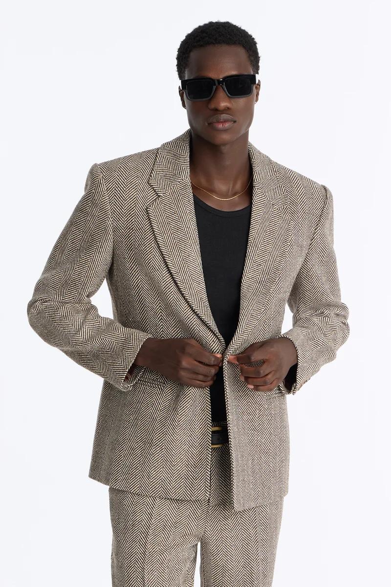Stylish man modeling a brown herringbone suit, buttoning the blazer, wearing black sunglasses. Showcasing versatile men's fashion.