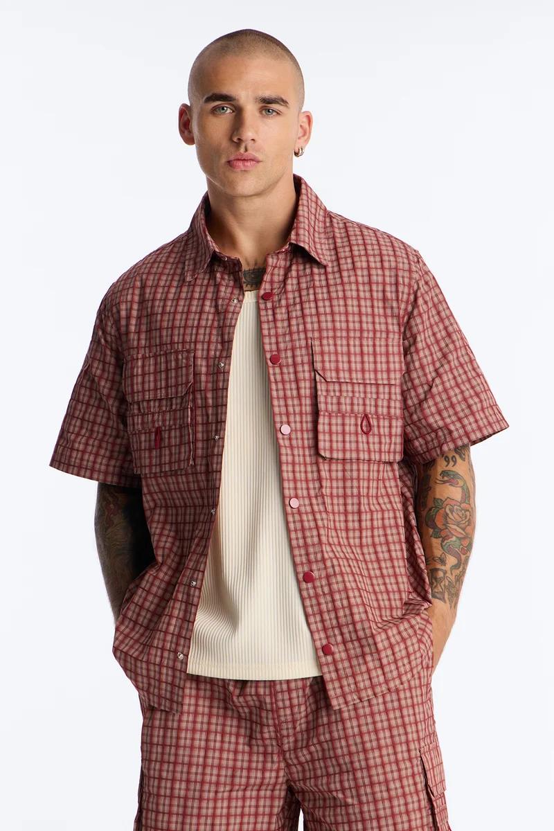 Man in a red and tan plaid short-sleeve shirt over a cream ribbed undershirt, paired with matching shorts, showcasing men's summer casual wear.