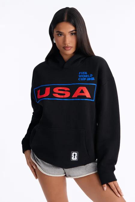 Woman in a black hoodie with red "USA" and blue outline, "FIFA WORLD CUP" in blue text, and grey shorts, posing frontally.