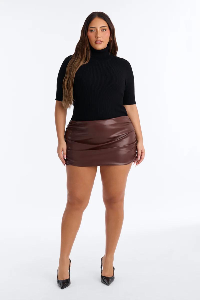 Modelo de talla grande con cuello alto negro y minifalda fruncida de cuero marrón, promocionando una nueva colección de moda de otoño.