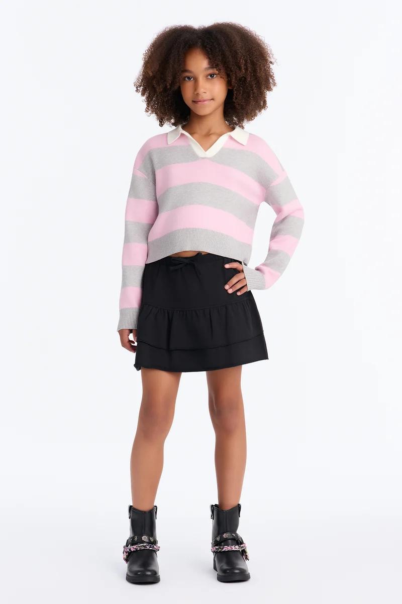 Niña con suéter de rayas rosa y gris con cuello, falda negra con volantes y botines.
