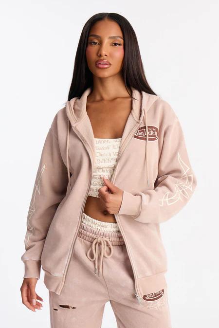 Mujer modela un conjunto Von Dutch rosa pálido: sudadera con cremallera y joggers rasgados. Mangas con gráficos blancos.