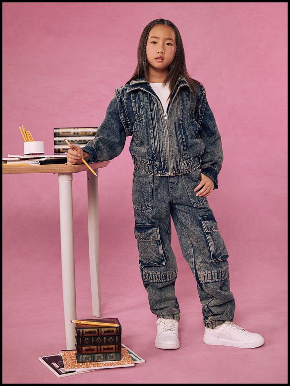 Girl in a blue kids denim jacket and cargo pants set stands near a school desk on a pink backdrop for a trendy back to school collection.
