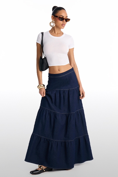 Woman models a white crop top and dark blue tiered denim maxi skirt with a black bag and sunglasses. Showcasing chic summer casual fashion.