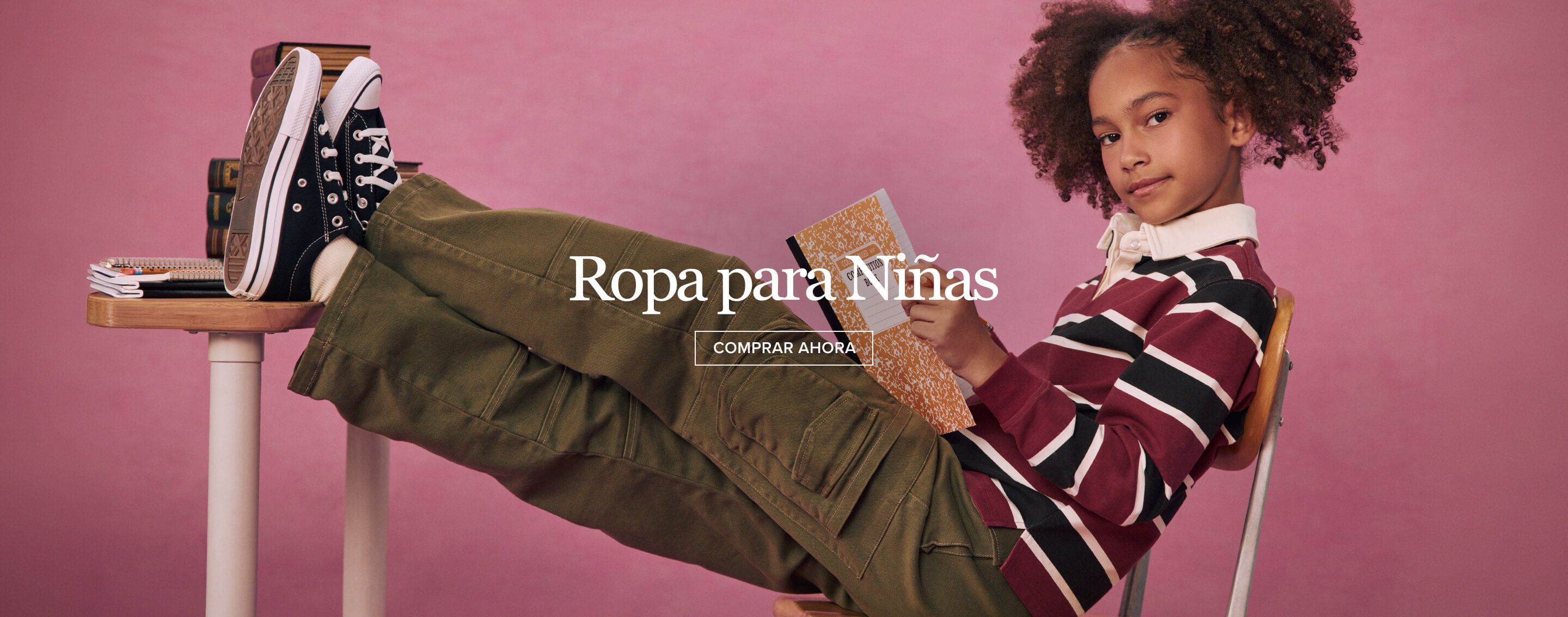 Niña que viste pantalones cargo verdes y polo a rayas posa al revés en un pupitre, sosteniendo una libreta sobre un fondo rosa.