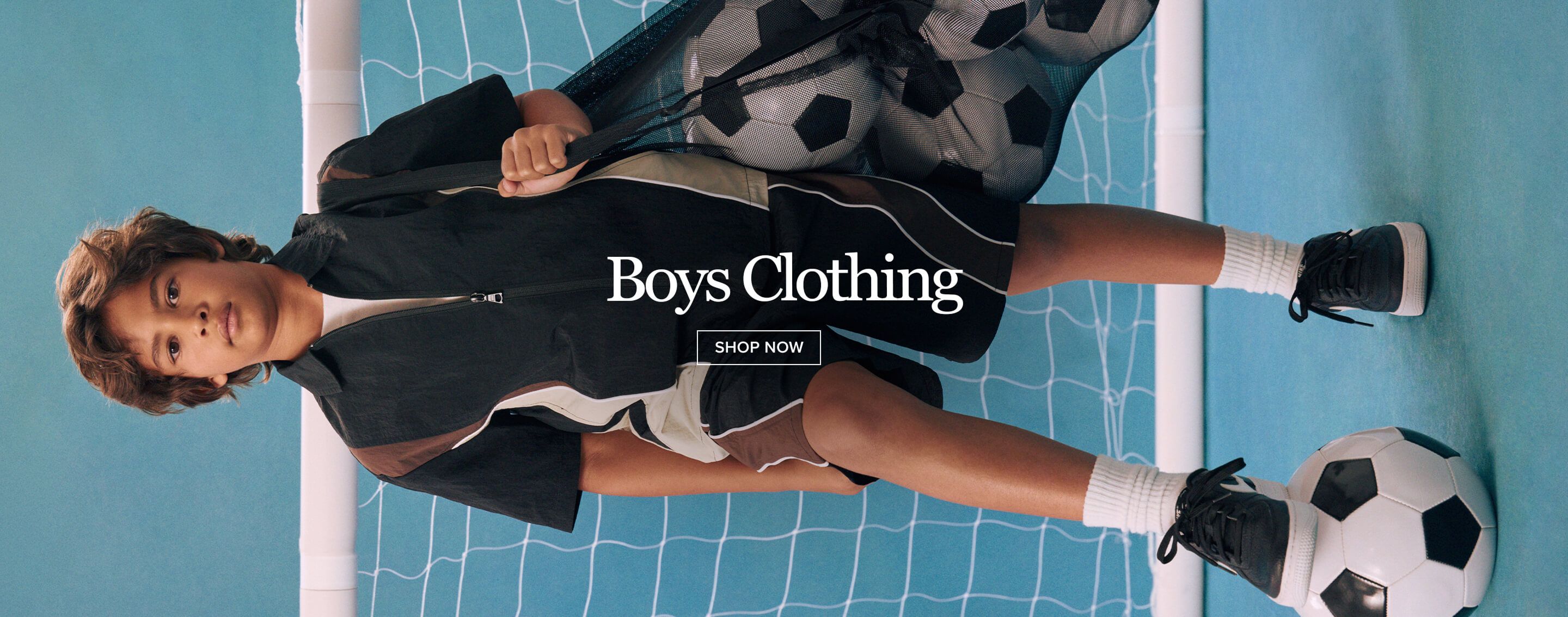 Young boy in black and brown activewear holds a bag of soccer balls against a blue net. Explore stylish boys clothing and sportswear now.