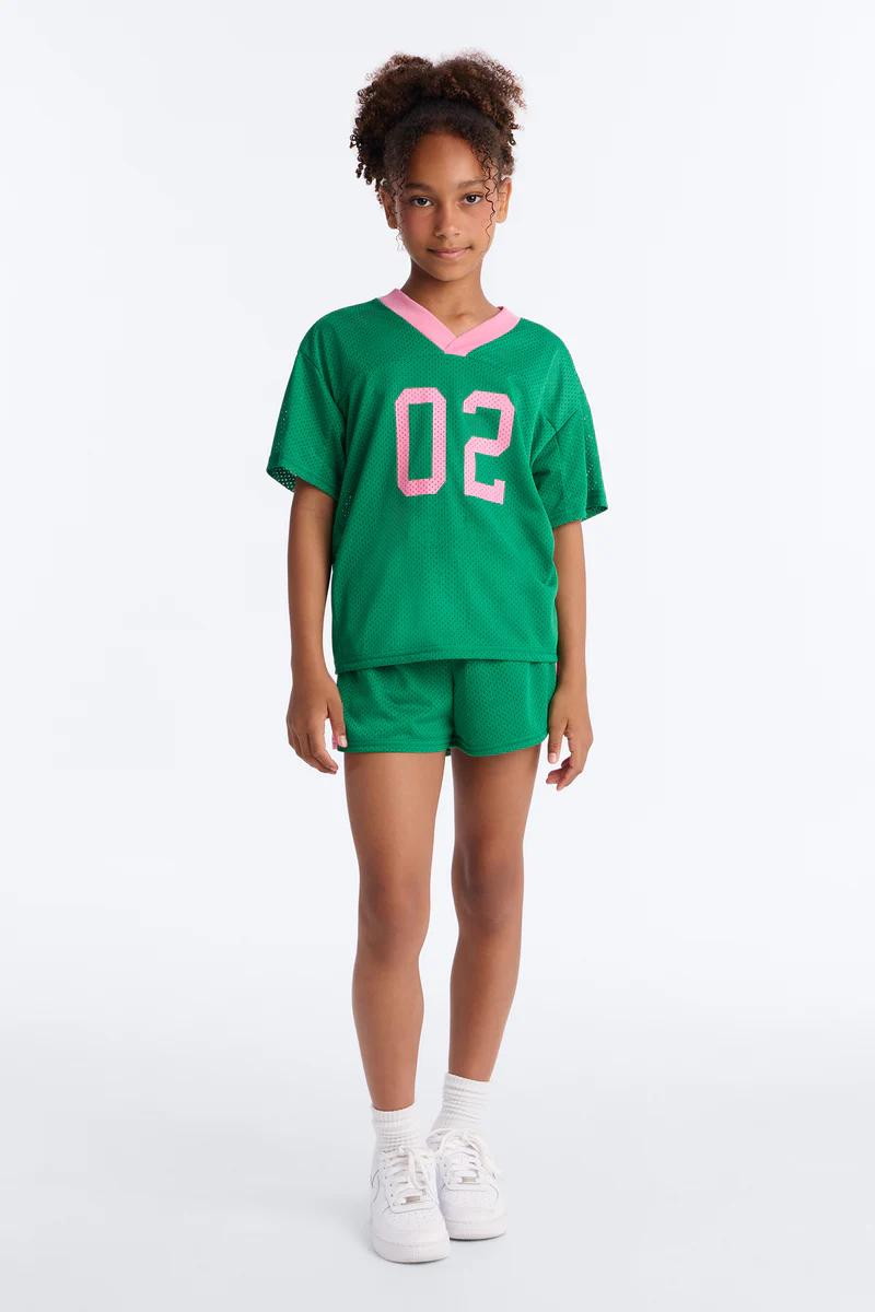 Niña con camiseta deportiva de malla verde (02) con ribete rosa y pantalones cortos a juego. Conjunto deportivo infantil.