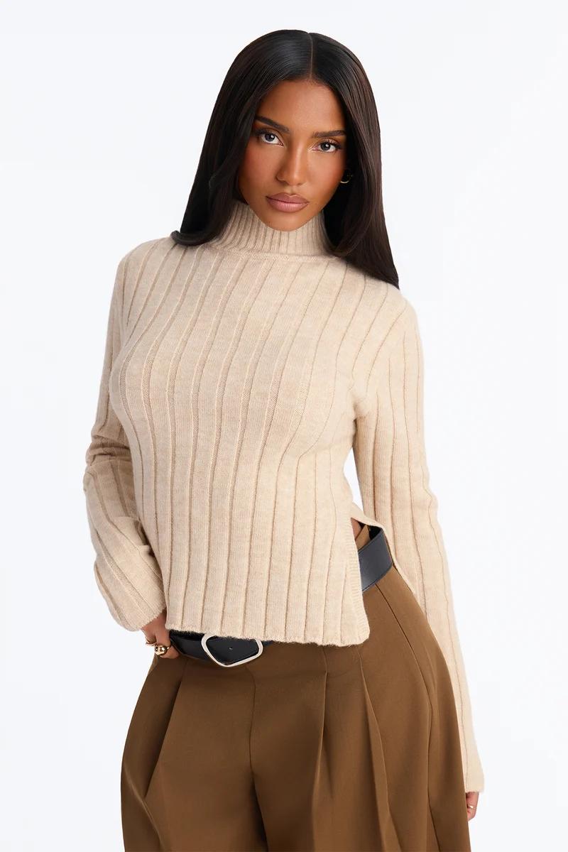 Woman wearing a beige ribbed knit turtleneck sweater with side slits and brown trousers.